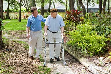 Younger man assisting an elderly man on a walk outside by a garden