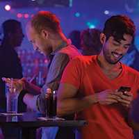 Foto de dos hombres en un club nocturno de pie y recostados hombro a hombro. Cada hombre mira hacia abajo en su teléfono celular mientras parecen enviar mensajes de texto.