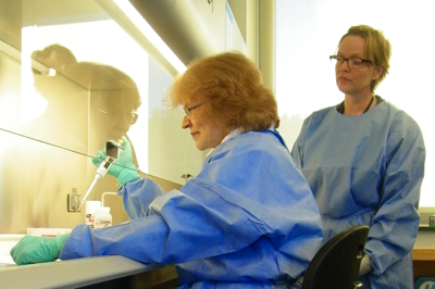 	CDC Microbiologists Olga Kosoy and Amy Lambert, in blue protective gear, working together in the lab. 