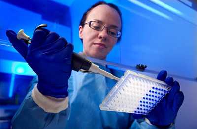Working in the confines of a ventilated hood, and wearing a blue-colored protective bio-hazard suite, this image depicts Centers for Disease Control (CDC) microbiologist Tatiana Travis, in the process of preparing a real-time polymerase chain reaction (PCR) test. In her right hand she holds a mechanized pipette containing a blue solution that she is pipetting into the 96-well plate held in her left hand.