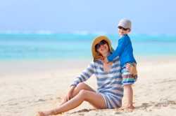 Photo of a mother and child on the beach. Both are wearing sunglasses, hats, and long-sleeved shirts.
