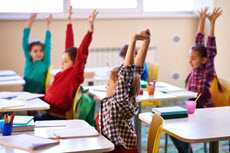 Kids sitting at their desks with both hands up in the air.