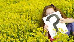 Girl siting in a meadow holding a sign with a question mark on it.
