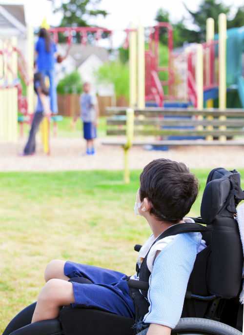 Disabled little boy in wheelchair watching children play on playground
