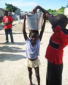 Photo of a girl with a bucket of water on her head and boy playing with it.