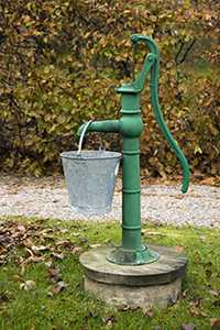 Image of a green water pump with a bucket hanging off of it.