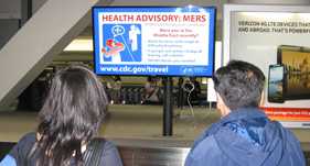 Man and woman at an airport reading a MERS-CoV health advisory on a TravAlert monitor