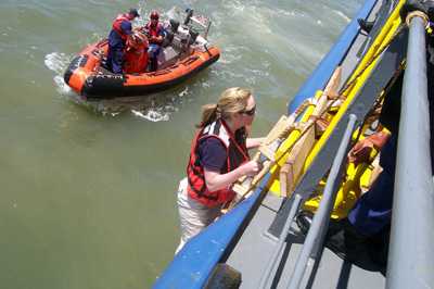 image of  a woman from the CDC using a Jacob’s ladder to conduct an offshore boarding of a vessel with US Coast Guard and Customs and Border Protection partners. A boat with two more people is seen in the background