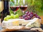 Assorted cheeses and grapes on a cutting board with two glasses of red wine in the background.