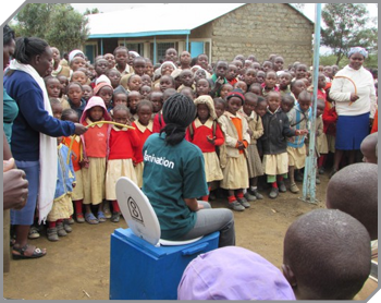 A Sanivation representative named Annette, in a green shirt, demonstrates a Blue Box toilet for a crowd of children and adults in a Kenyan village