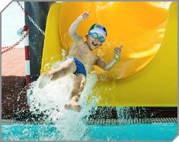 Boy giving two thumbs up as he flies off the end of a yellow waterslide in a big splash of water.