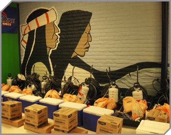 Image of equipment grouped into sets (boxes, orange bags, coolers and a pump spray bottle commonly used for bug treatment) in front of a brick wall painted with a mural  - the profiles of two American Indians