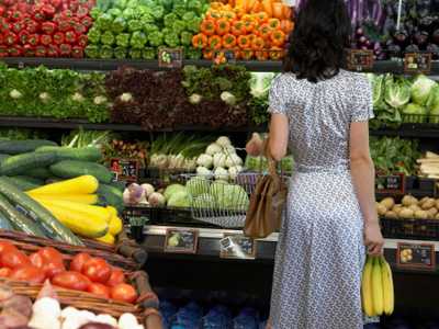 Brunette woman, in a white and black patterned dress, holds a bunch of bananas and a shopping basket as she considers a wide arrange of colorful fruit and veggies in a grocery store.