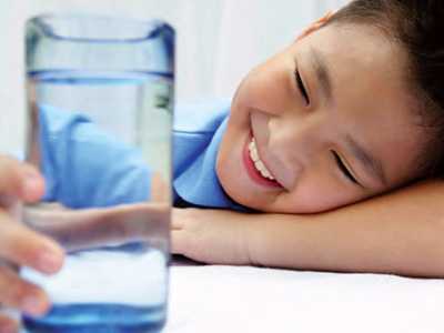 A young boy in a blue shirt sits at a white table, laying his head on his arm, smiling at a clear glass of water in his hand.