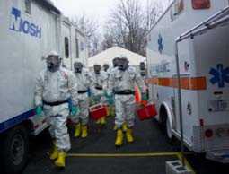 a group of seven emergency responders in protective gear walking toward the viewer between an ambulance on the right and a NIOSH branded trailer on the left.