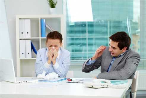 woman sneezing into a tissue next to a male co-worker hiding his face