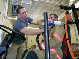 	An assistant digitizes a firefighter study participants wrist location using a Faro Arm device, while the participant reaches for his seatbelt in turnout gear.