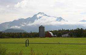 large green field with silo and barn in the background, in front of a cloud capped mountain range.