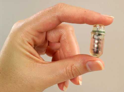 The SmartPill, approximately 1.06 inches long by .46 inches in diameter, held by a person’s thumb and index finger.
