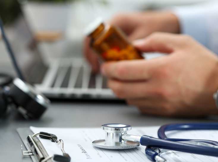 Foto de las manos de un médico sosteniendo un frasco de medicamento recetado, con una computadora, un estetoscopio y un bloc de recetas médicas al fondo.
