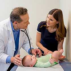 Male doctor with a stethoscope listens to a child’s heartbeat, as the mother talks to doctor.