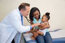 Un médico escucha el latido del corazón de un niño por medio de un estetoscopio mientras la madre sostiene al niño en sus brazos. 
